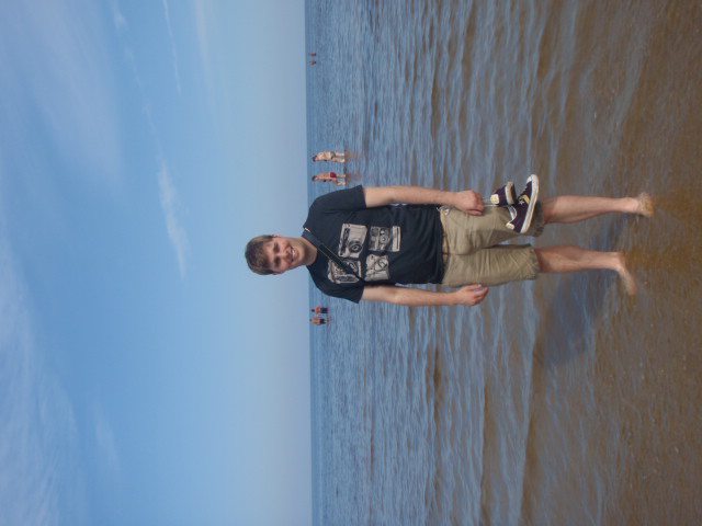 Guy standing in the surf at the beach. He is holding his shoes in his left hand.